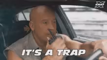 A bald man wearing a sleeveless tan buttondown (Vin Diesel in a Fast & Furious movie) driving a car and speaking into a walkie talkie. The words It's a trap are on the bottom in a white overlay.